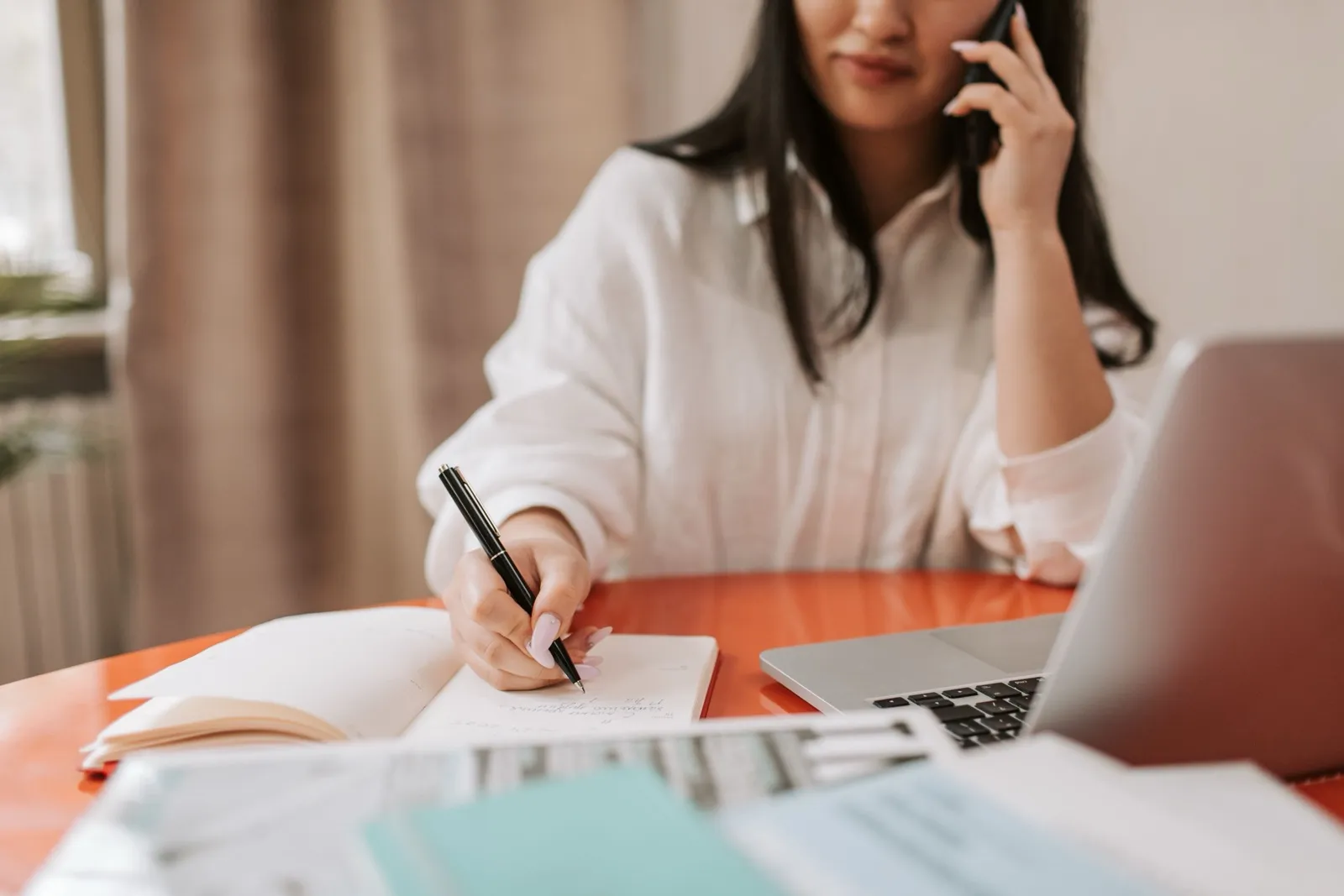 オフィスで会社員が電話を耳に当てながらメモを取り、机の上に書類が積まれている様子