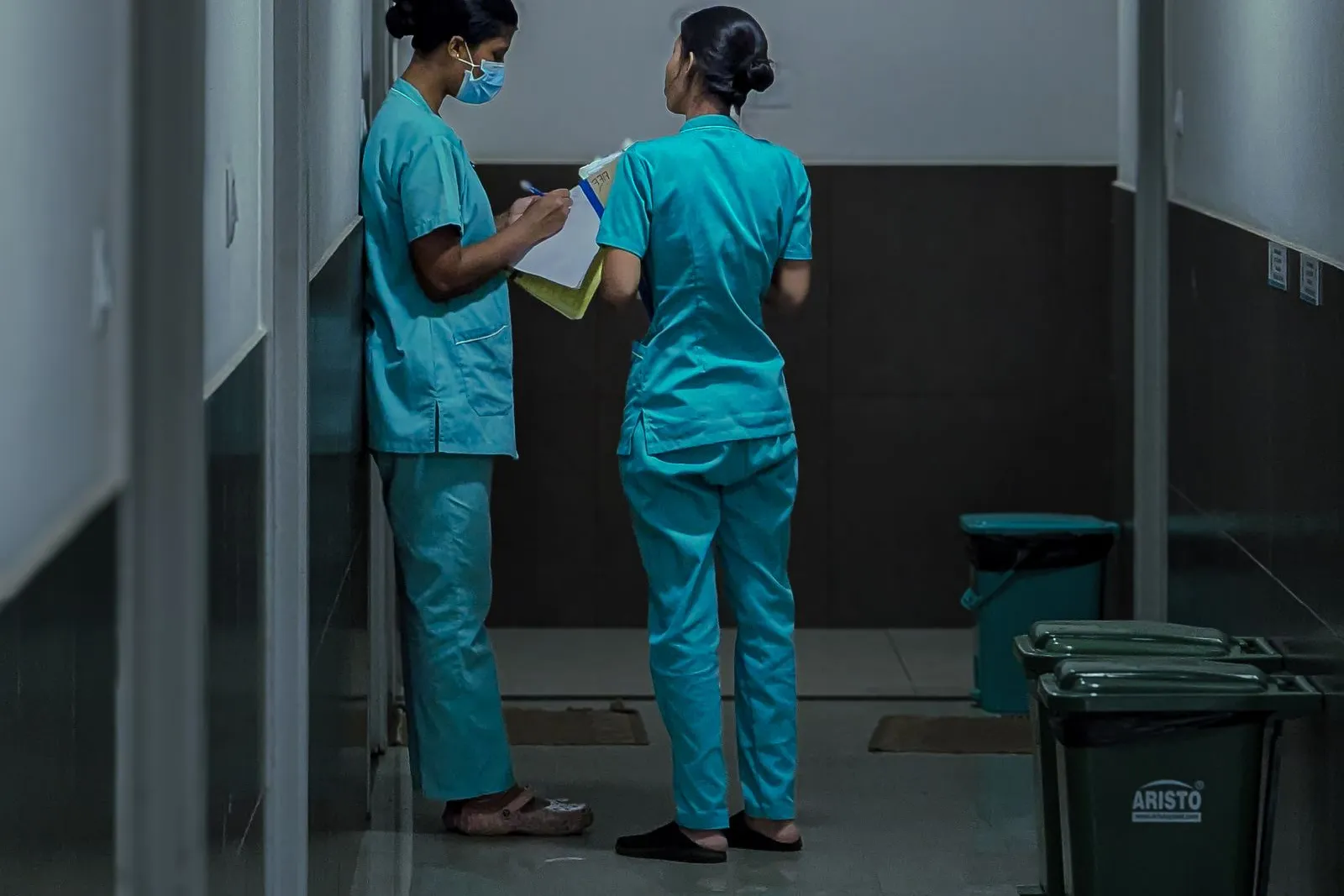 薄暗い廊下で、クリップボードの用紙を確認しながら話し合う2人の医療・介護スタッフの後ろ姿